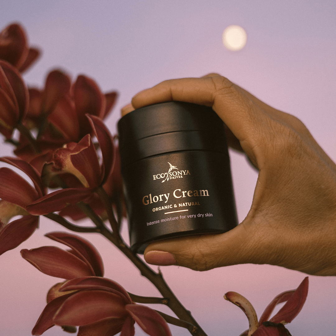 Hand holding a jar of Eco by Sonya Glory Cream with flowers in the background - Eco By Sonya UK